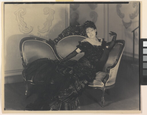 A black-and-white photograph of a White woman in a long, formal gown and elbow-length gloves posing on a sofa.