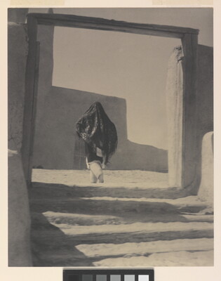 A black-and-white photograph of a person wearing a long shawl over their head and torso while walking through the wood gate of an adobe building.