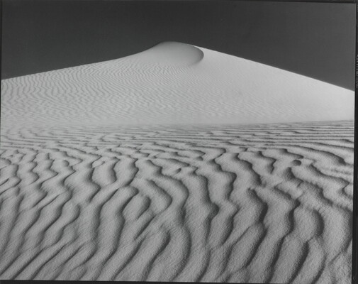 A sepia-toned photograph of a deeply-rippled sand dune rising to a point into the sky.