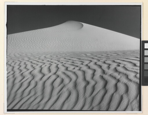A sepia-toned photograph of a deeply-rippled sand dune rising to a point into the sky.