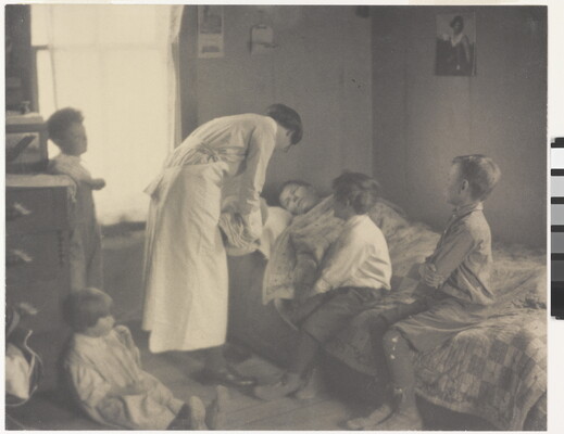 A black-and-white photograph of a nurse bending over a person in a bed, two children sitting on the bed, and two children behind the nurse.