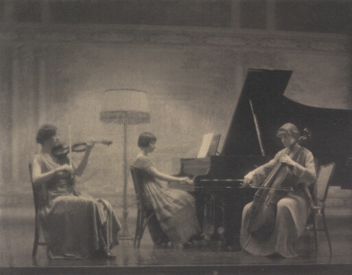 A black-and-white soft-focus photograph of three seated White women: one plays a violin, one a piano, and the third a cello.