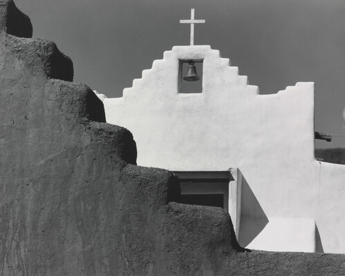 A black-and-white photograph of a terraced adobe bell tower with a cross on top, partially hidden by a terraced adobe wall.