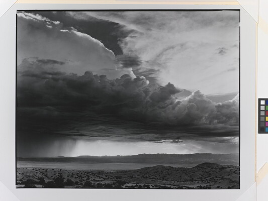 A black-and-white photograph of torrential rain and heavy storm clouds passing into a brightly lit plain.