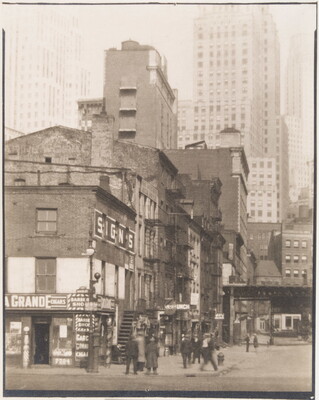 A black-and-white photograph of people on a street corner near three- and four-story buildings with skyscrapers in the background.