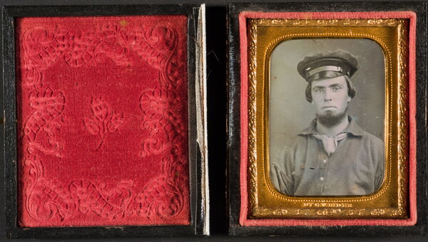 An open photograph case showing, on the right, a black-and-white photograph in a gold frame of a young man posed in a military hat.
