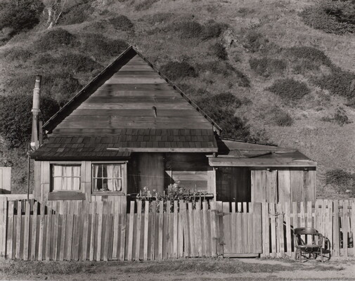 A black-and-white photograph of a small clapboard house behind a picket fence with a chair next to the gate.