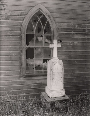 A black-and-white photograph of a tall, white tombstone with a cross on top next to a wood building with a broken lancet-shaped window.