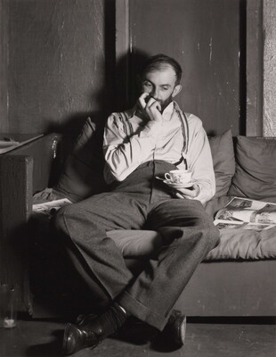 A black-and-white portrait photograph of a bearded White man seated on a sofa with one hand up to his mouth, the other holding a teacup over his lap.