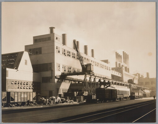 A black-and-white photograph of a multi-story industrial building with multiple smokestacks on top and tracks for a streetcar in front.
