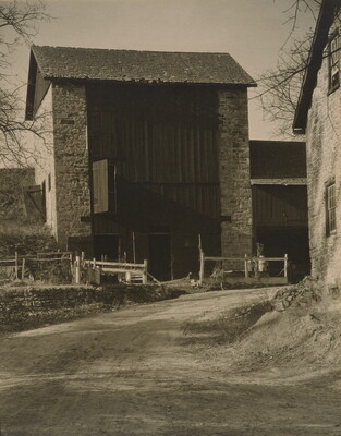 A black-and-white photograph of a dirt road leading to a multi-story stone-sided structure with a wood facade.
