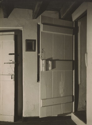 A black-and-white photograph of an interior with light-colored plaster walls and two wood doors: one mostly closed to the left, and the one on the right open.