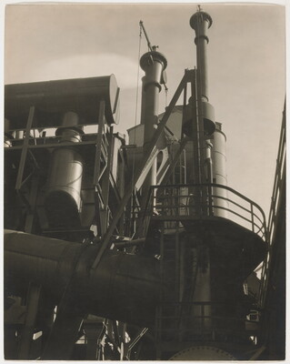 A black-and-white photograph of the exterior of a factory taken from the ground looking up toward the top of its stacks.