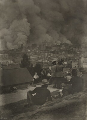 A black-and-white photograph of a group of people on a hillside watching smoke rise from a city skyline in the distance.