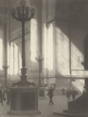 A black-and-white photograph of people walking through a sunlit plaza with towering light posts and columns.