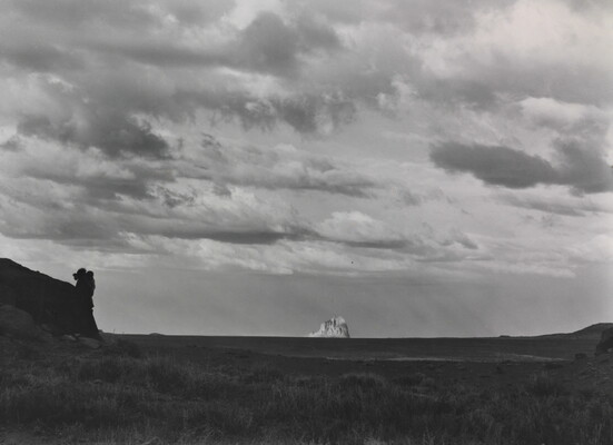A black-and-white photograph of an open field with a large, white rock formation in the distance under a cloudy sky.