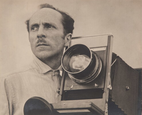 A sepia-toned portrait photograph of a balding White man with a mustache beside a large-format camera.