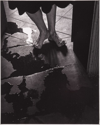 A black-and-white photograph of a person's bare feet, toes stretched up off the tile floor to avoid puddles.