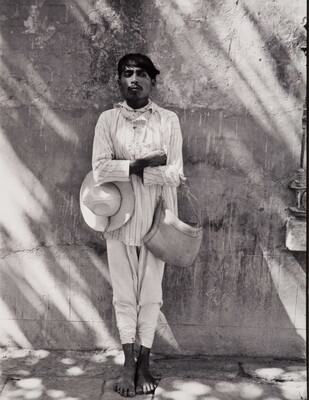A black-and-white photograph of a dark-skinned man in light-colored shirt and pants, holding a hat under one crossed arm and a bag on the other, leaning against a wall.