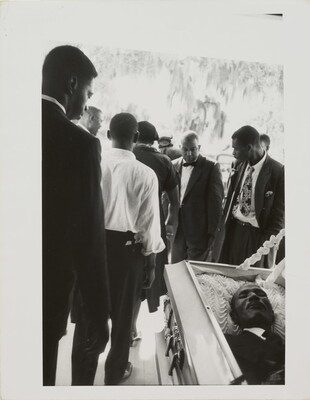 A black-and-white photograph of Black men in suits filing by an open casket.