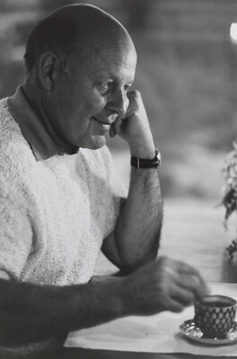 A black-and-white portrait photograph of a bald White man in profile resting his chin on a closed fist as his other hand stirs liquid in a cup.