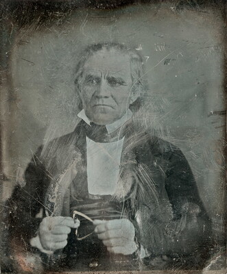 A worn and scratched black-and-white photograph of a seated, older White man in a suit.