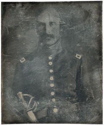 A dark, heavily scratched black-and-white photograph of a White man with a moustache, wearing military regalia and a sword.