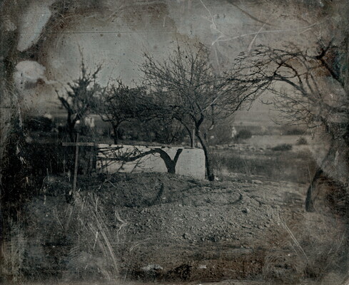 A scratched black-and-white photograph of a wooden cross marking a burial site, surrounded by leafless trees on a dirt hill.