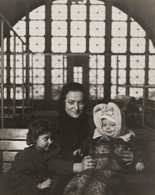 A black-and-white photograph of a White woman with two young children seated on a bench, with a large arched window behind them.