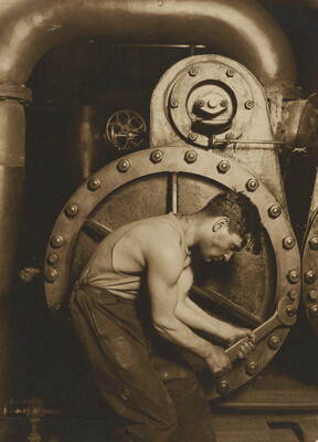 A black-and-white photograph of a muscular man crouched next to a large piece of machinery that he is adjusting with a large wrench.