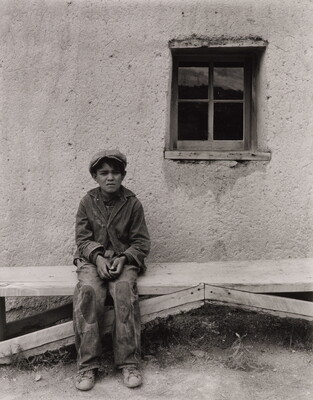 A black-and-white photograph of a squinting medium-skinned boy wearing a cap, jacket, and pants with patches on the knees, seated on a wooden bench in front of an adobe building.