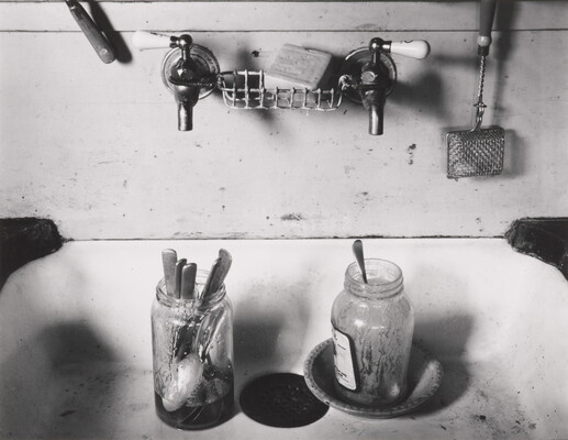 A black-and-white photograph of a two-tap sink with two dirty mason jars full of spoons.