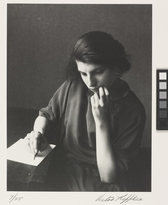 A black-and-white photograph of a young White woman seated at a table writing on a piece of paper.