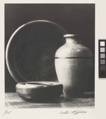 A black-and-white photograph of a short wood bowl, a large pear-shaped vase, and a standing plate behind them.