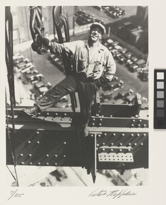 A black-and-white photograph of a smiling White man standing on a beam holding onto a chain, wearing a newsboy cap, cars in parking lots far below.