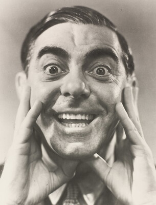 A black-and-white portrait photograph of a White man, hair slicked back, hands framing his cheeks, mouth open in a wide smile.