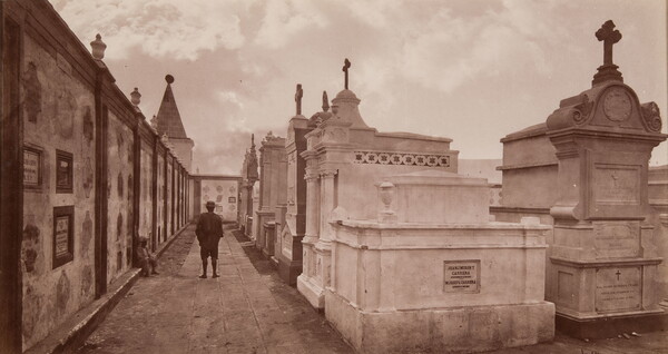 A sepia-toned photograph of a man standing among a row of large marble mausoleums, burial vaults, and cremation niches.