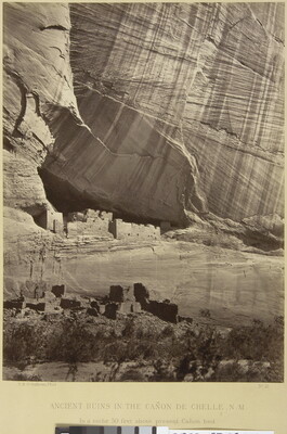 A sepia-toned photograph of cliff dwellings at the base of a large, sheer cliff.