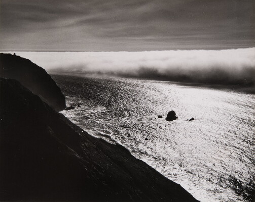 A black-and-white photograph taken from a hillside overlooking a calm ocean as thick clouds roll in or out.