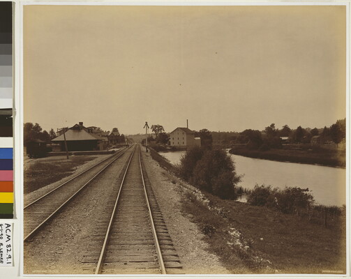 A sepia-toned photograph of railroad tracks leading to several buildings, and water to the right of the tracks.