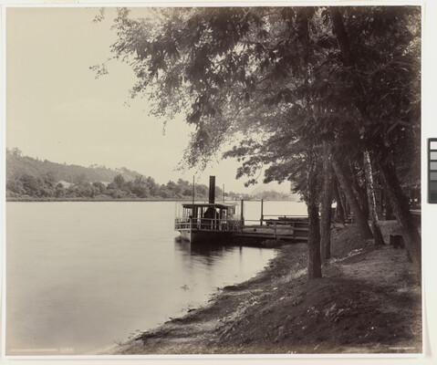 A black-and-white photograph of a boat docked on a river and trees on the shore.