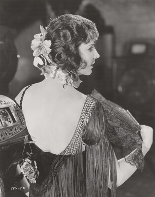 A black-and-white photograph, taken from the back, of a White woman wearing a low-back fringed frock, and fluttering a lacy fan.
