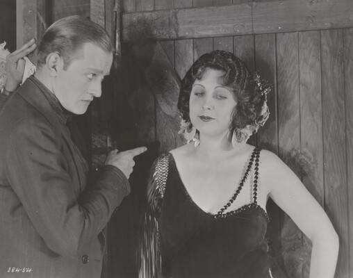 A black-and-white photograph of a young White man looking sternly and pointing at a young White woman as she gazes off to the side with a slight smile.