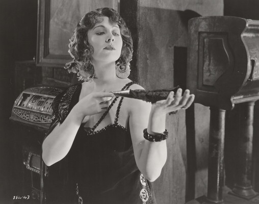 A black-and-white photograph of a White woman wearing a 1920s-style dress holding a feathered fan in her outstretched hands.