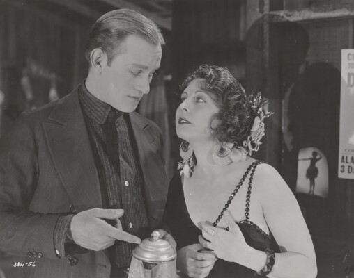 A black-and-white photograph of a White man and woman dressed in 1920s attire looking at each other as the man points to a coffee pot he is holding.