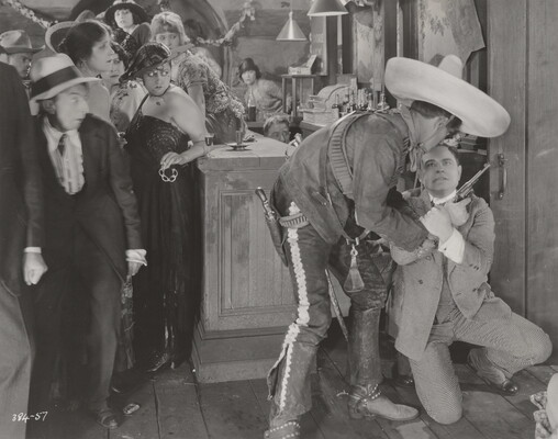 A black-and-white photograph of a group of people in a bar watching two men, one wearing a large sombrero and holding a revolver, wrestle over the gun.