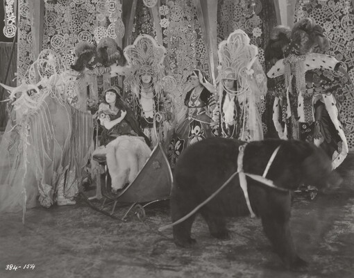 A black-and-white photograph of group of people on a stage wearing elaborate costumes, one of whom sits in a sleigh harnessed to a bear, in front of a set depicting snowflakes and icicles.
