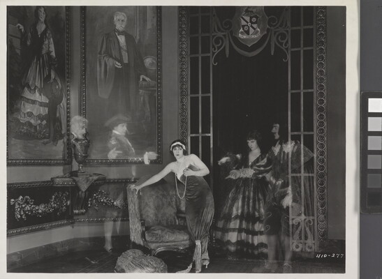 A black-and-white photograph of a White woman standing next to a loveseat in a lavish room as four transparent people in historical costumes point to her.