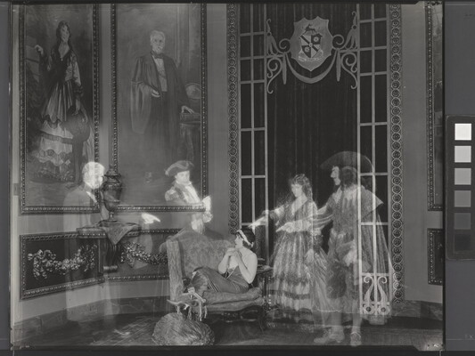 A black-and-white photograph of a White woman huddled on a loveseat in a lavish room as four transparent people in historical costumes point to her.