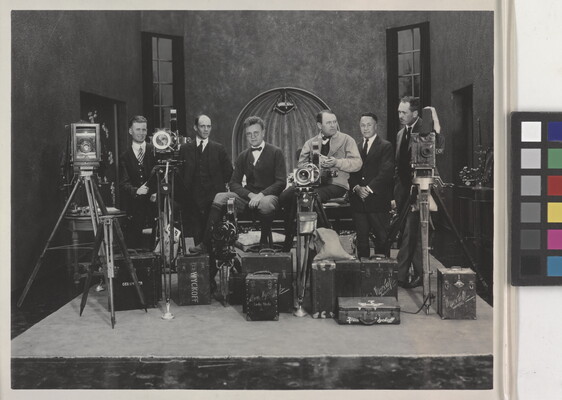 A black-and-white photograph of a group of White men in suits on a movie set posed with multiple cameras and lighting equipment.